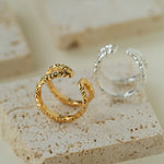 Two textured snake-shaped open rings, one gold and one silver, displayed on beige stone blocks.